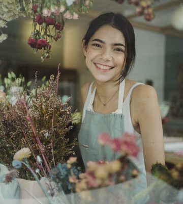 Through window of charming female worker in apron standing among flowers in store smiling at camera