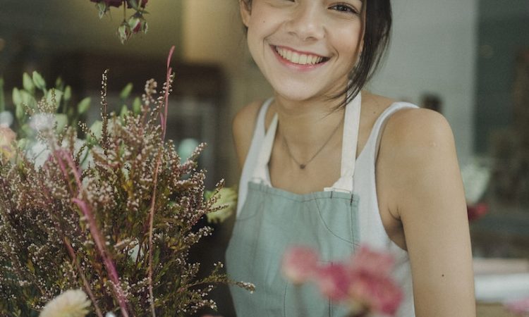 Through window of charming female worker in apron standing among flowers in store smiling at camera